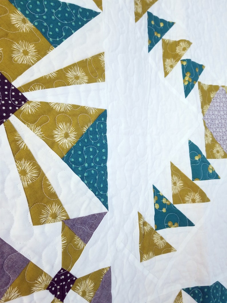 May include: A quilt with a geometric pattern in shades of teal, gold, purple, and white. The quilt is made of fabric with a variety of patterns, including polka dots, floral designs, and geometric shapes.