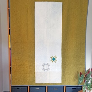 May include: A white quilt with two blue and green star designs is hanging on a wooden shelf with black storage bins underneath. The quilt is flanked by two panels of mustard yellow fabric.