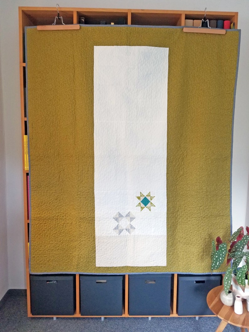 May include: A white quilt with two blue and green star designs is hanging on a wooden shelf with black storage bins underneath. The quilt is flanked by two panels of mustard yellow fabric.
