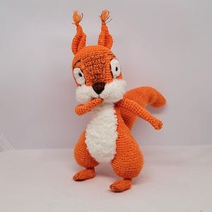 Peut inclure: Une peluche d'écureuil au crochet, orange et blanc, avec un nez marron et de grands yeux. L'écureuil a une poitrine blanche duveteuse et une grande queue. Il est posé avec une patte près de sa bouche, lui donnant une expression curieuse.