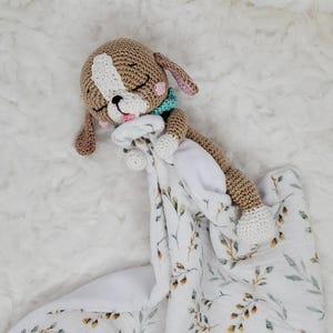 May include: A crocheted puppy dog toy with closed eyes, a white patch on its head, and pink accents. The puppy rests on a white and floral-patterned blanket. The blanket has a soft, textured appearance, and the toy has a teal scarf.