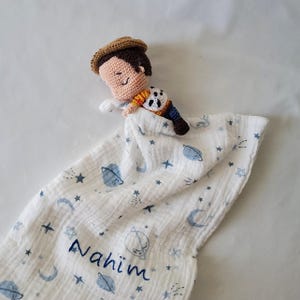 Könnte beinhalten: Eine gehäkelte Schnuffeltuch-Decke mit einer kleinen Stofffigur, die einen braunen Hut, beigefarbene Haut und ein weiß-blau-gelbes Outfit trägt. Der weiße Musselinstoff ist mit blauen Planeten, Sternen und Monden bedruckt und mit dem Namen Nahim bestickt.