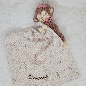 Può includere: Un giocattolo a forma di scimmia all'uncinetto con un fiocco rosa e una coperta. La coperta presenta un motivo leopardato nei toni del beige e del marrone, con il nome "Emma" ricamato sopra.