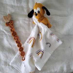 Peut inclure: Un jouet pour chien au crochet jaune et noir est posé sur une couverture en mousseline blanche avec des motifs arc-en-ciel aux couleurs douces. Une attache-tétine en bois en forme de renard et une attache-tétine en crochet marron sont également présentes.