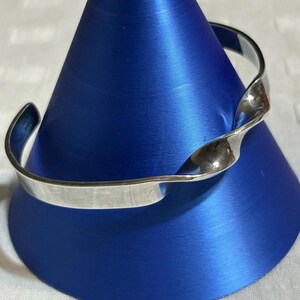 May include: A silver-toned cuff bracelet featuring a smooth, flat band that transitions into a twisted, spiraled section. The metal has a polished, reflective finish, and the open-ended design allows for adjustment around the wrist.