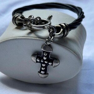 May include: Black twisted cord bracelet with a silver-tone cross pendant. The cross has a detailed design with rounded edges and small dot accents. The bracelet features a hook clasp and is displayed on a white surface.