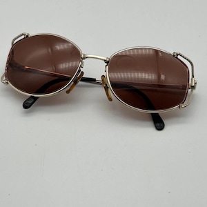 May include: Vintage sunglasses with oval brown lenses and a silver metal frame. The frame features decorative detailing on the temples and a bridge. The arms are black with brown ear pieces. The sunglasses are set against a white background.