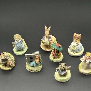 May include: A collection of eight small, hand-painted animal figurines featuring anthropomorphic characters including rabbits, cats, and a fox. Each figure is depicted in various poses, such as reading, holding items, or wearing clothing, set on individual circular bases.