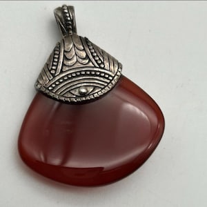 May include: A pendant featuring a polished, teardrop-shaped, reddish-brown stone. The stone is capped with an ornate, silver-colored metal piece with decorative patterns. The pendant is designed to be worn as jewelry.