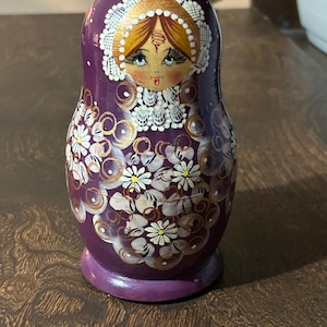 May include: A purple Matryoshka doll with a painted face and floral design. The doll features a traditional Russian design with a white lace headdress and gold accents. The body is decorated with white flowers and gold swirls.