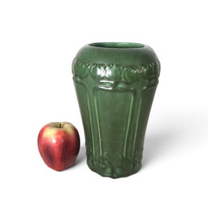 Puede incluir: Un jarrón de cerámica verde bosque mate con forma cilíndrica cónica y un patrón floral en relieve en los bordes superior e inferior. Una manzana roja y amarilla está colocada junto al jarrón para proporcionar una referencia de escala.