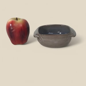 May include: A small, square, gray ceramic bowl with two handles. The bowl has a blue glaze on the inside and a brown glaze on the outside. The bowl is sitting on a white surface.