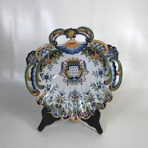 May include: A white ceramic shell-shaped dish with blue, yellow, and green floral designs. The dish features a coat of arms with a crown and fleur-de-lis.
