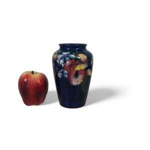 Puede incluir: Un jarrón de cerámica azul oscuro con un diseño floral con flores rojas, amarillas y blancas. El jarrón tiene un cuerpo redondeado y un borde ligeramente acampanado. Hay una manzana al lado del jarrón.