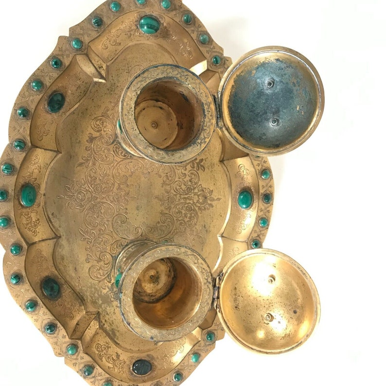 May include: Gold-toned inkwell set with three compartments, each with a lid. The inkwell is decorated with an intricate floral pattern and has green gemstones around the edge.