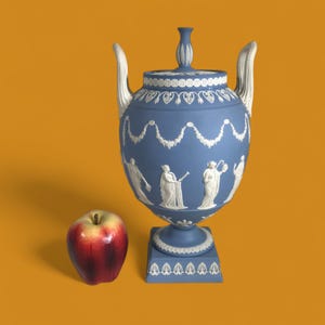 Może przedstawiać: Niebiesko-biała ceramiczna urna z pokrywką, uchwytami i kwadratową podstawą. Urna posiada białe figury reliefowe i dekoracyjne girlandy. Czerwono-żółte jabłko leży obok urny.