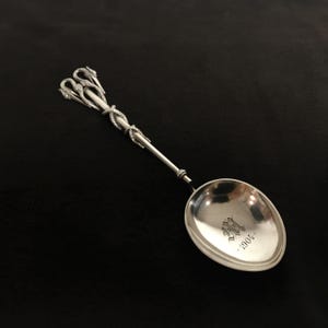 Può includere: Un cucchiaio d'argento con un manico decorativo che presenta tre teste di cigno intrecciate. La ciotola del cucchiaio è incisa con le iniziali "M.H." e la data "1904."