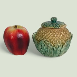 以下が含まれることがあります： 緑色の蓋とパイナップルのデザインが施された陶器の壺。壺は緑の葉で飾られており、表面はテクスチャ加工されています。背景には赤いリンゴがあります。