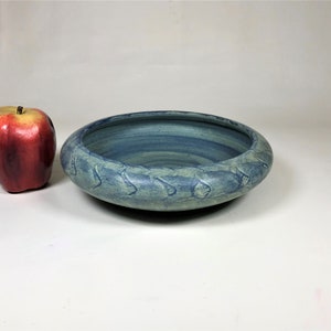 May include: A handmade, blue ceramic bowl with a textured rim. The bowl has a wide, shallow design and a gradient of blue hues. A red apple is next to the bowl.