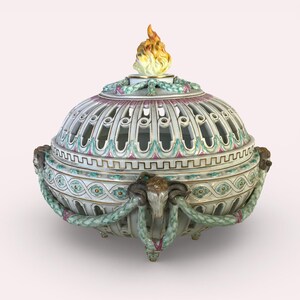 May include: A decorative, ornate ceramic urn with a domed lid and a flame finial. The urn features a white base with intricate cut-out designs, accented with pink, green, and gold details. Ram's head handles and garland add to the classical aesthetic.