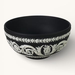 Può includere: Una ciotola in ceramica nera con un design in rilievo bianco e ornato attorno al bordo. La ciotola ha una finitura opaca e una forma semplice ed elegante. Il motivo decorativo comprende motivi floreali e fogliari.