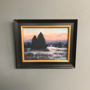 Pode incluir: Pintura emoldurada de uma paisagem de inverno com um pôr do sol rosa e laranja no fundo. Três árvores verde-escuras ficam em primeiro plano, com um campo nevado à sua frente.