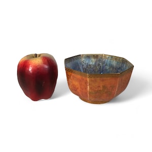 May include: A small, octagonal ceramic bowl with a blue and gold glaze. The bowl has a textured surface and is sitting on a white background.