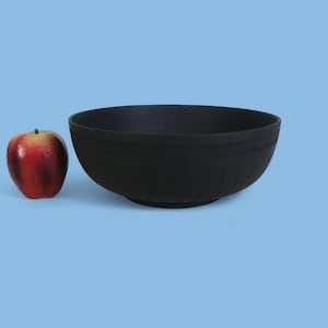 May include: A large, black bowl with a textured exterior sits next to a red apple on a light blue background. The bowl is round and has a wide opening. The apple is red with yellow accents.