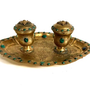 May include: A gold-toned inkwell set with two inkwells and a tray. The inkwells are decorated with green gemstones and have ornate tops. The tray has a rope-like handle on each side and is decorated with green gemstones around the edge.