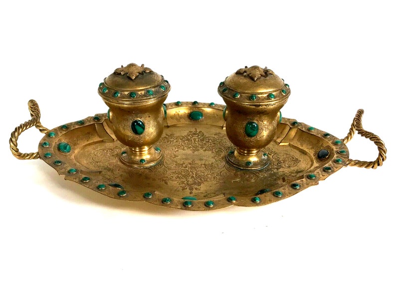 May include: A gold-toned inkwell set with two inkwells and a tray. The inkwells are decorated with green gemstones and have ornate tops. The tray has a rope-like handle on each side and is decorated with green gemstones around the edge.