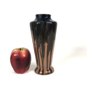 Pode incluir: Um vaso de cerâmica alto com a parte superior azul escuro e gotas marrons sobre uma base bege. O vaso tem uma forma alargada e uma base decorativa. Uma maçã vermelha está ao lado do vaso.