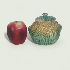Può includere: Una mela rossa accanto a un vaso decorativo in ceramica con coperchio. Il vaso è rotondo con una superficie strutturata, con un motivo a losanghe e foglie. Il vaso è principalmente verde e marrone.