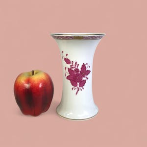 May include: A white porcelain vase with a flared top and a floral design in shades of magenta. The vase has gold trim around the top and base. An apple is next to the vase.