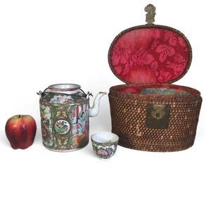 Può includere: Un servizio da tè antico con una teiera decorativa, una tazza e un cestino intrecciato con interno floreale rosso. Una mela rossa è a sinistra del servizio da tè. La teiera e la tazza hanno disegni floreali e figurativi colorati.