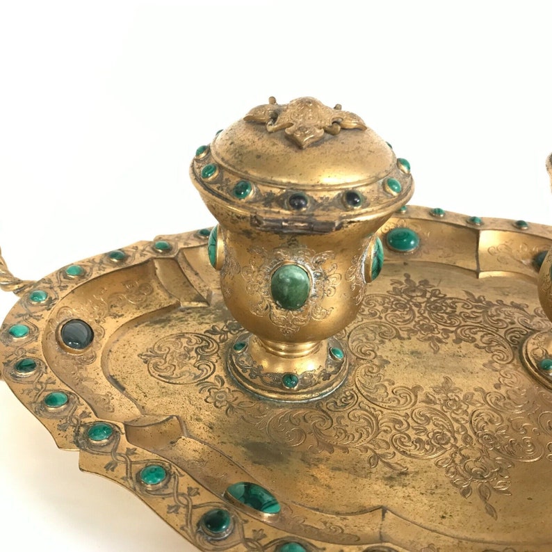 May include: A gold inkwell set with two inkwells and a tray. The inkwells are decorated with green gemstones and intricate floral patterns. The tray is also decorated with green gemstones and floral patterns.