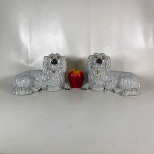 May include: Pair of white porcelain dog figurines with black eyes and gold accents. The dogs are lying down with their heads turned to the side.