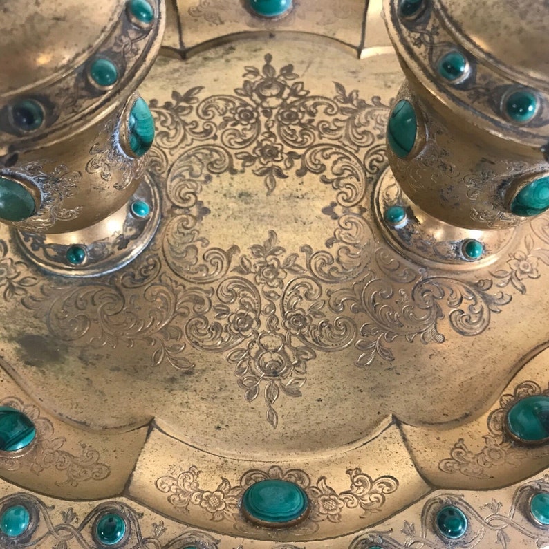 May include: A gold tray with intricate engraved designs and green gemstone accents. The tray has a scalloped edge and is decorated with floral motifs.