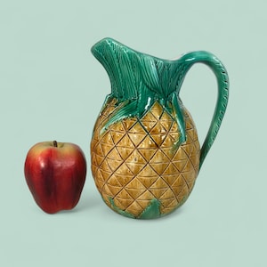 May include: A ceramic pineapple-shaped pitcher with a green leafy top and handle. The pitcher is tan with a diamond pattern. A red apple sits next to the pitcher. The background is a light green color.