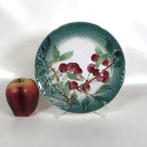 Pode incluir: Um prato de cerâmica branca com uma borda verde e branca e um desenho em relevo de um ramo de cerejeira com cerejas vermelhas.