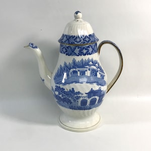 Puede incluir: Una tetera de porcelana blanca con un diseño azul y dorado. La tetera tiene un cuerpo redondo con un pico acampanado y una tapa con un pomo. El diseño presenta un paisaje con árboles, un edificio y un puente.