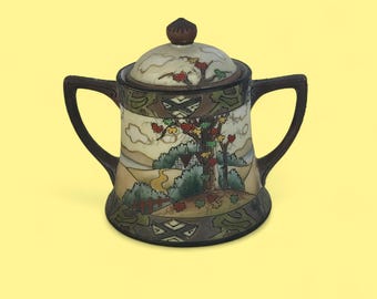 May include: A ceramic sugar bowl with a lid, decorated with a landscape scene of a tree, house, and hills. It has two handles and a dark brown trim. The lid is topped with a small knob. The bowl is likely made of ceramic.