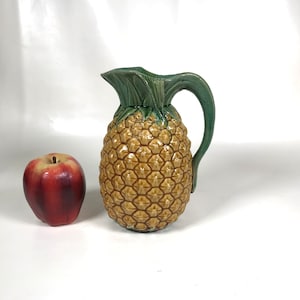 May include: A ceramic pitcher shaped like a pineapple, with a green handle and leafy top. The pitcher is tan with a textured surface, and sits next to a red apple. The pitcher is a decorative item.