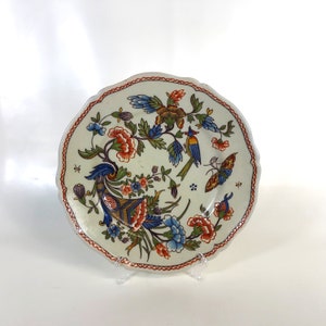 May include: A white ceramic plate with a scalloped edge features a colourful floral design with birds and a butterfly. The design includes blue, red, orange, yellow, and green colours.