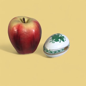 May include: A red and yellow apple next to a white egg-shaped trinket box with green floral designs. The trinket box has a gold band around the middle and green trim. The apple is a medium size.
