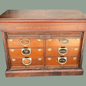 May include: A vintage wooden cabinet letter file with six drawers. The cabinet is brown with brass hardware and labels. The top of the cabinet reads "AMBERG'S CABINET LETTER FILE". The labels on the drawers include "Bills Payable", "Present Year", "Miscellaneous", and "Past Year".