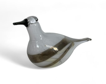 May include: A white glass bird with a black beak and gold and white stripes. The bird is sitting on a white surface.
