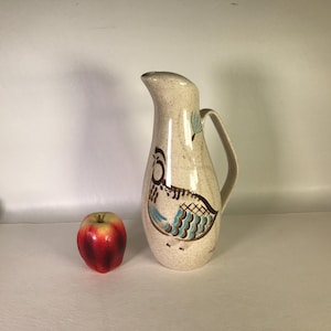 May include: A tall, speckled beige ceramic pitcher featuring a stylized brown and blue bird illustration on the front, accompanied by a single red apple placed to its left, showcasing a curved handle and a tapered spout for pouring liquids.