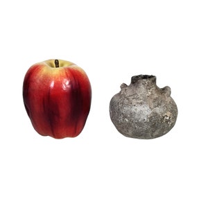 May include: A red apple next to a small, gray, textured, round vase with two small handles. The apple is a vibrant red with yellow accents. The vase has a rough, aged appearance.