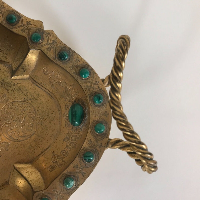 May include: A gold tray with a rope handle and green gemstone accents. The tray has an intricate design and is perfect for serving or displaying items.