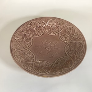 Puede incluir: Un plato grande, redondo, de cerámica rosa con un diseño en relieve de hojas y enredaderas alrededor del borde. El centro del plato presenta un diseño en relieve de una escena con dos figuras.
